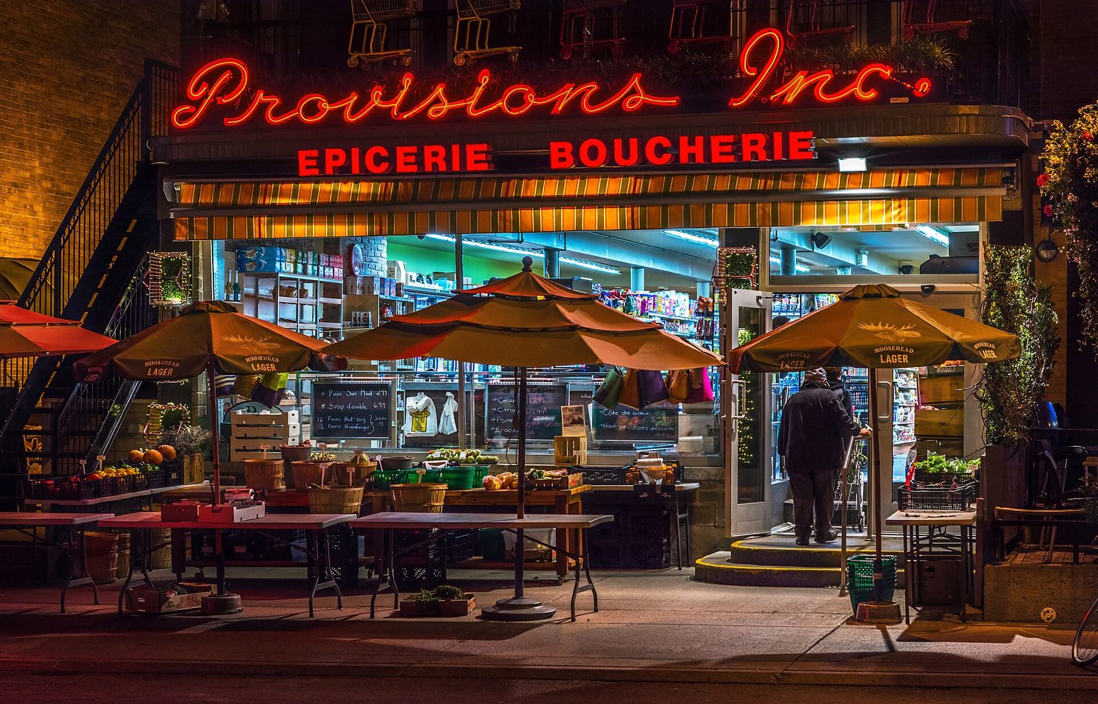 A lit grocery and butcher shop front at night with produce under parasols