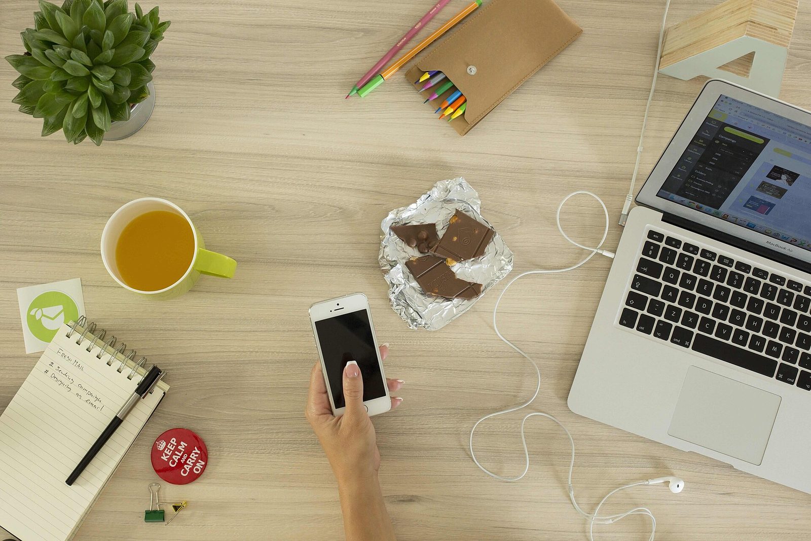 A hand holding a smartphone over a desk with a laptop, a notebook and pencils